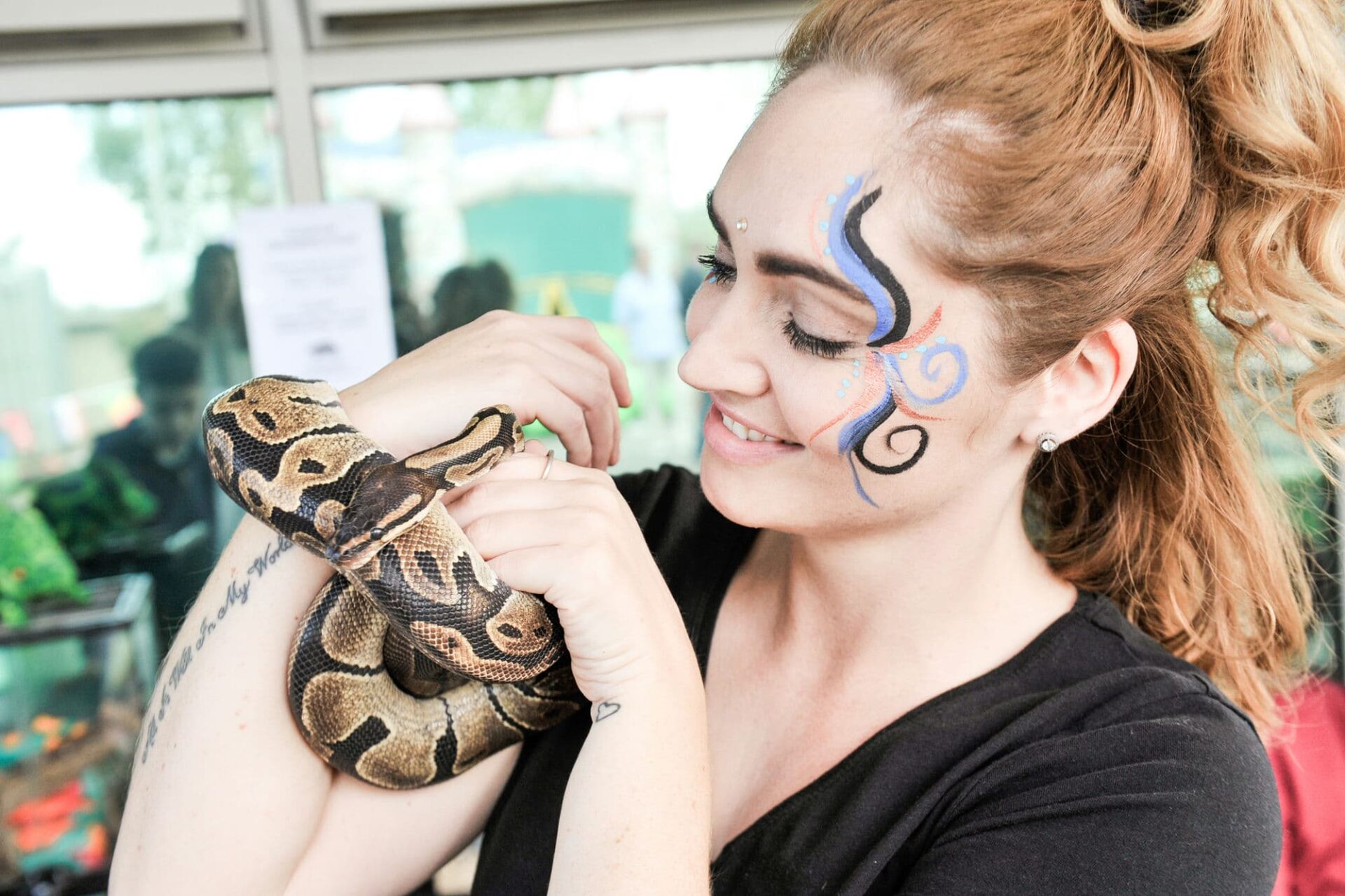 An attractive young woman is holding a constrictor snake and smiling. Her face is painted