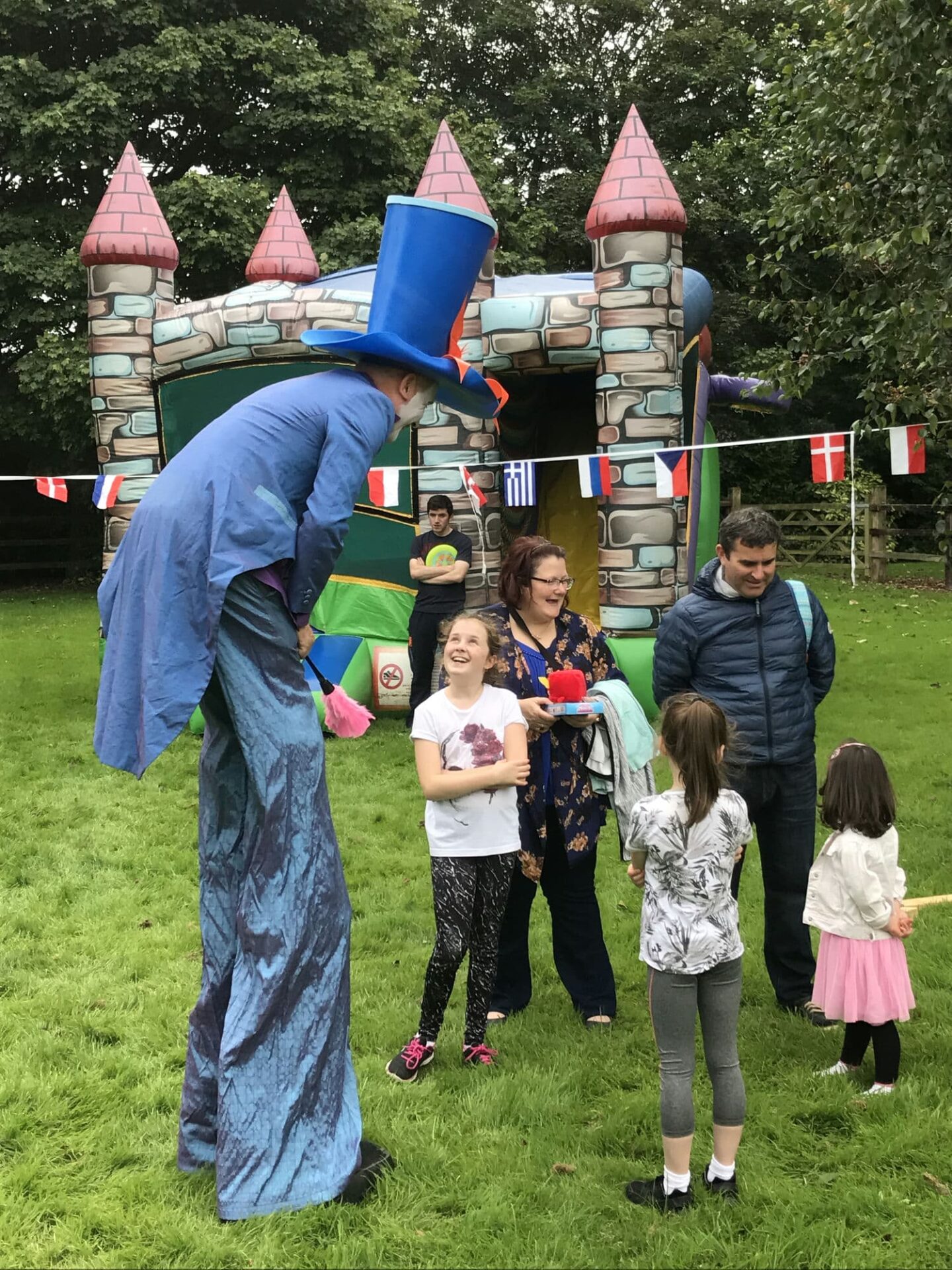 A family at a "family fun day" summer party is entertained by a stilt walker man in a blue top hat and fancy dress. There is an inflatable bouncy castle in the background